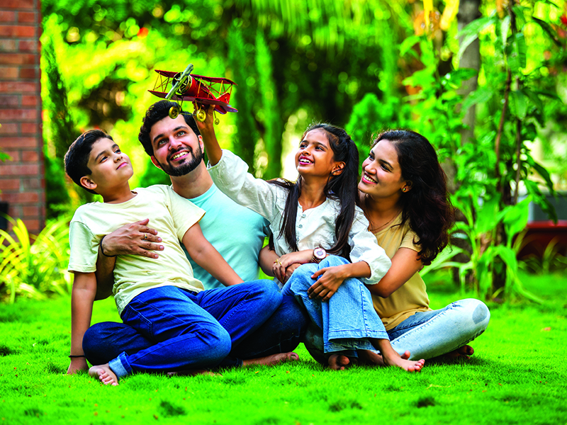 Joyful Indian couple with their kids sitting in a park, embracing each other and admiring a toy plane held by their daughter, symbolizing love, family time, imagination, and togetherness, aspiration