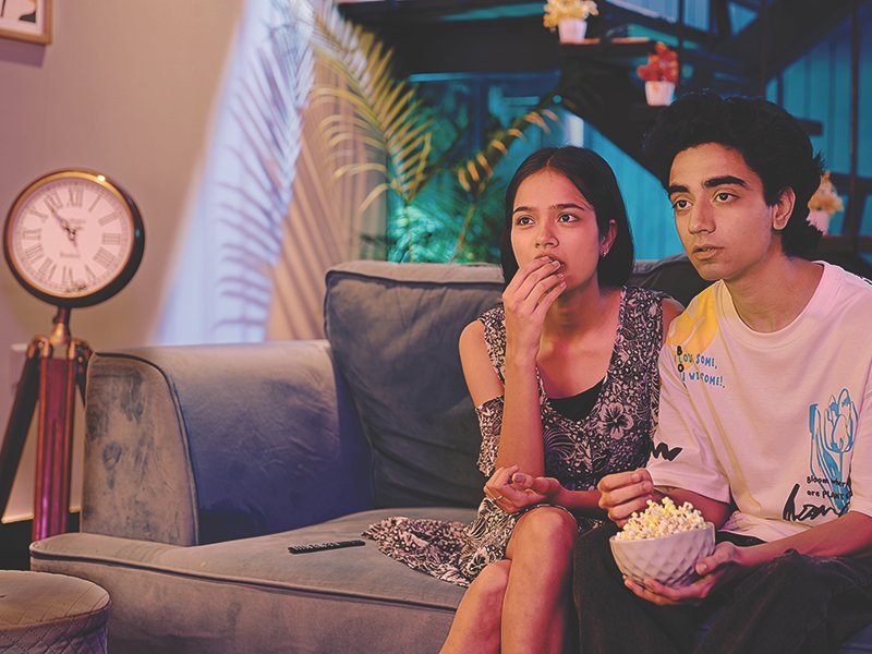 Two terrified Indian boyfriend with girl sitting on sofa watching horror film TV at home late night spend time together. Beautiful teen friend couple excited eat popcorn snack looking action video